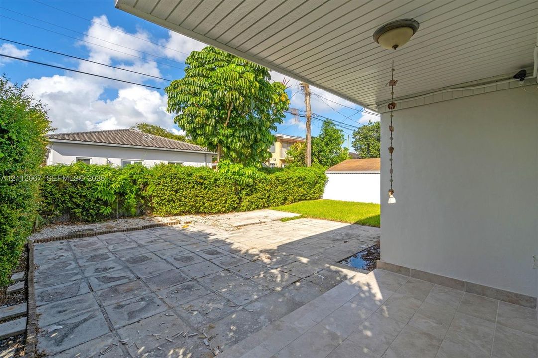 Covered patio with tile flooring and ceiling-mounted light fixture, featuring a stone-paver rear terrace, manicured hedge perimeter, and dedicated lawn area