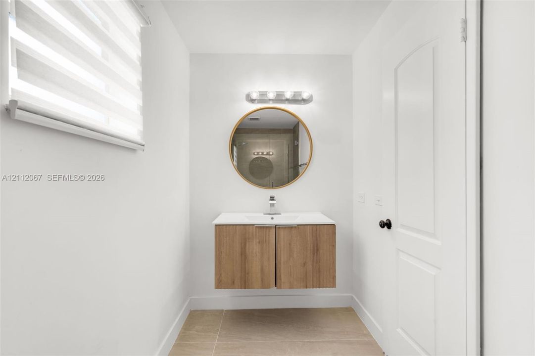 Floating vanity with wood-finish cabinetry, solid surface countertop, circular mirror with metallic frame, four-light vanity fixture, and patterned window shade