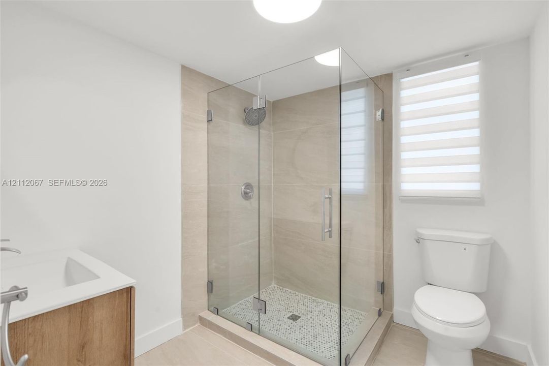 Walk-in shower with a frameless glass enclosure, neutral-toned wall tile, pebble-style floor tiling, wood-finish vanity, and integrated sink