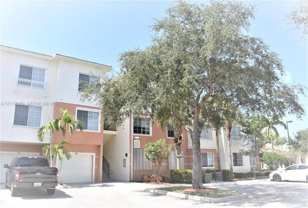 Multi-story residential building featuring a stucco exterior, integrated garage, private balconies with railings, and mature landscaping