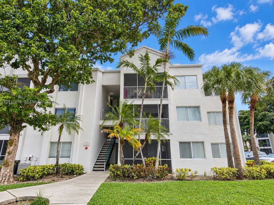 Multi-story stucco exterior featuring screened balconies, metal railed staircase, mature landscaping, and palm trees