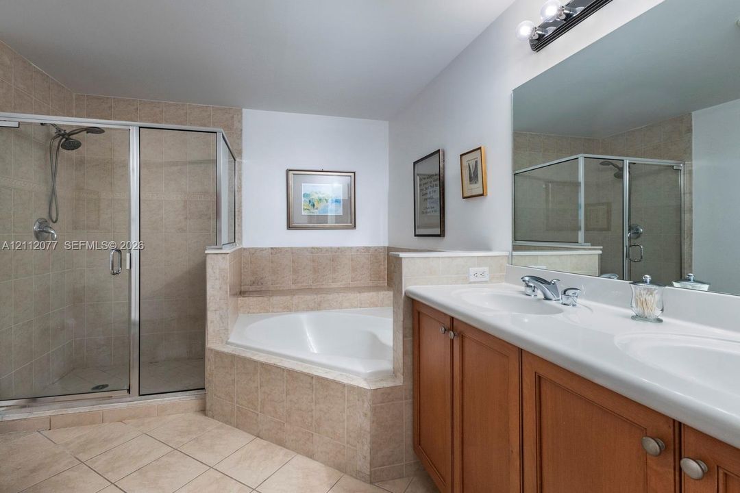 Bathroom featuring a dual-sink vanity with a solid-surface countertop, corner soaking tub with tile surround, glass-enclosed shower, and tile flooring