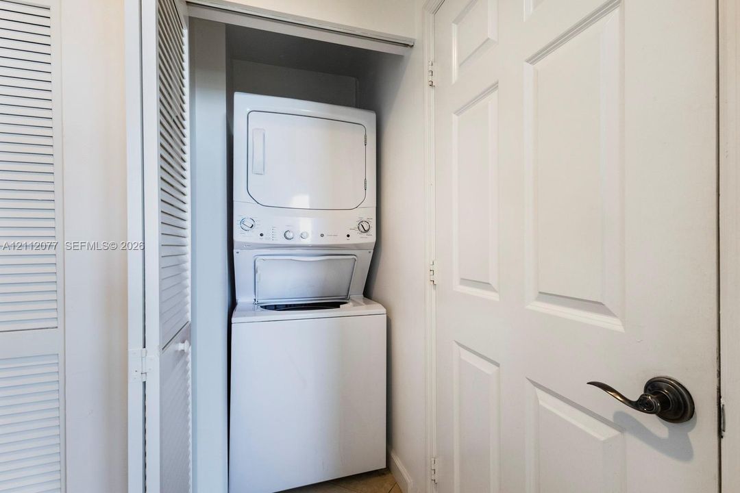 Stacked washer and dryer unit housed in a closet with white louvered doors and a traditional panel door with an oil-rubbed bronze lever handle