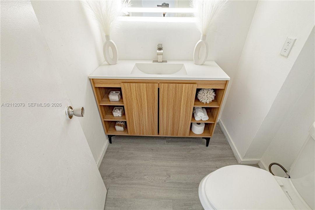 Modern vanity featuring wood-finish cabinetry, open shelving, a clean-lined sink basin, white countertop, and an integrated LED-backlit mirror