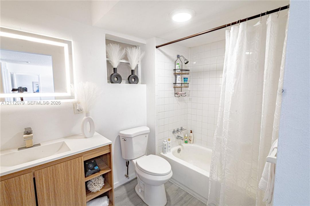 Bathroom featuring a wood-finish vanity with an integrated basin, LED-backlit mirror, shower-tub combination with white wall tile, and recessed shelving