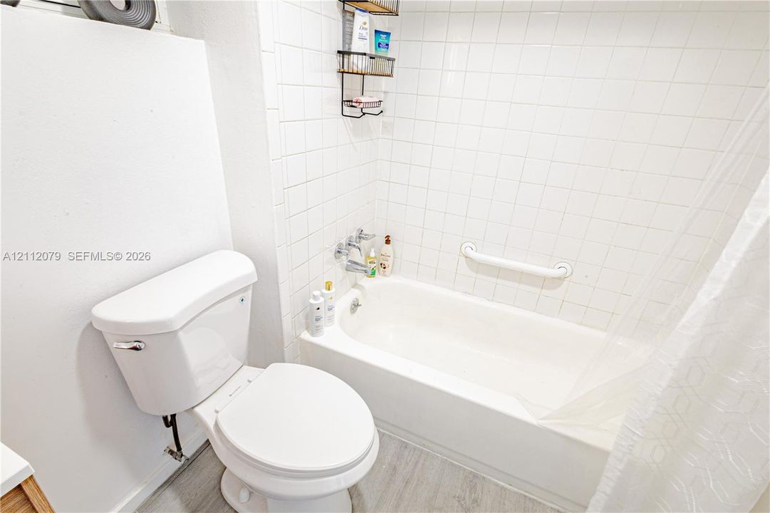 Bathroom featuring a porcelain tub, wall-mounted safety grab bar, square tile surround, wood-finish flooring, and a standard-height toilet