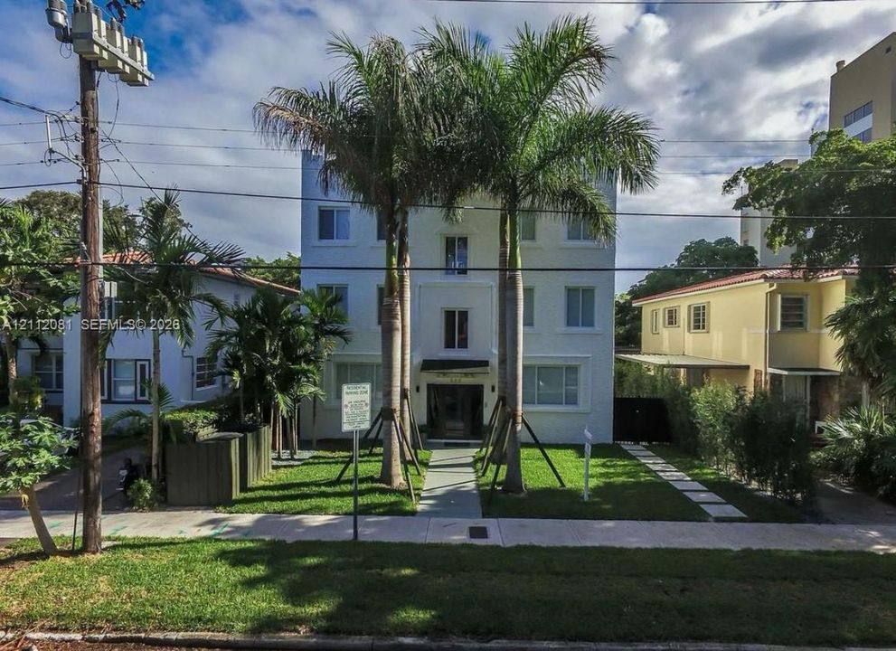 Three-story multi-unit structure featuring a white facade, symmetrical window placement, a paved walkway, manicured lawn, and mature palm trees