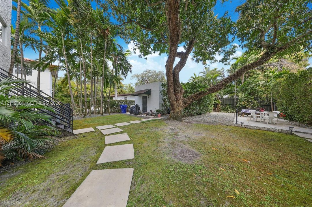 Private backyard featuring a paved stone walkway, established mature trees, low-maintenance landscaping, and a concrete patio area