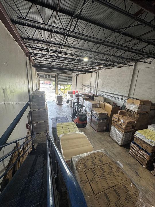 Warehouse interior featuring an open-span ceiling with exposed steel trusses, painted masonry walls, concrete flooring, and metal staircase access