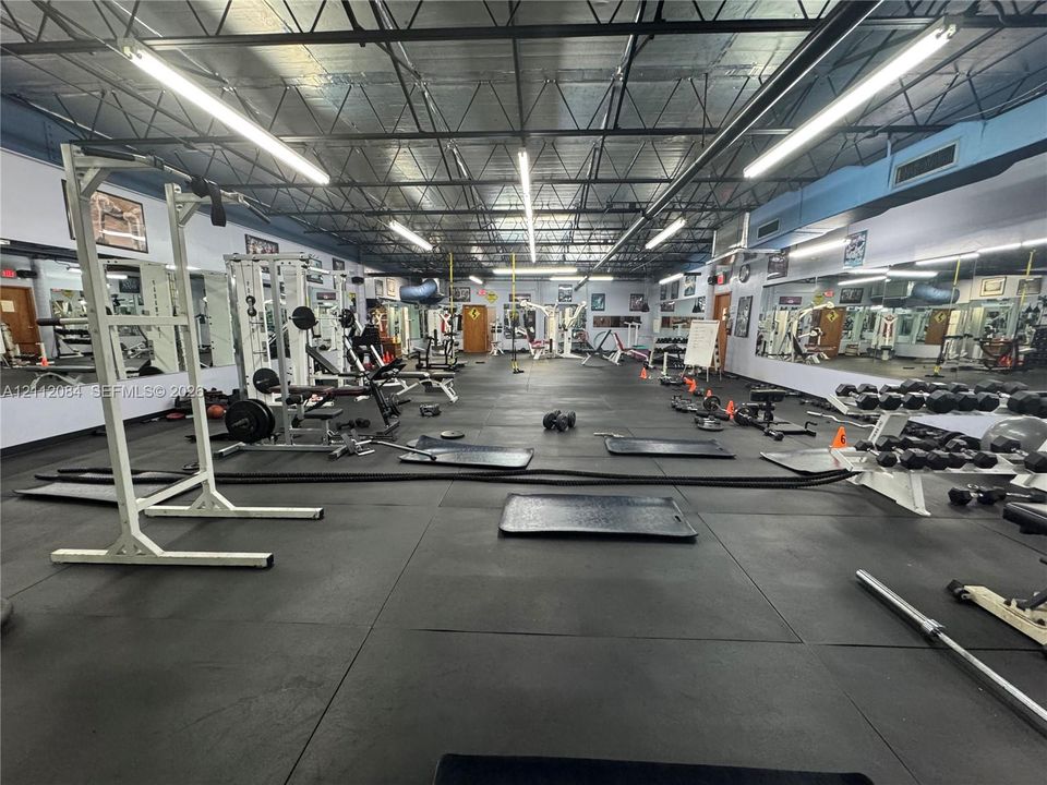 Open-concept room featuring exposed steel truss ceiling, industrial-style suspended linear lighting, extensive wall-to-wall mirrors, and impact-resistant rubber flooring