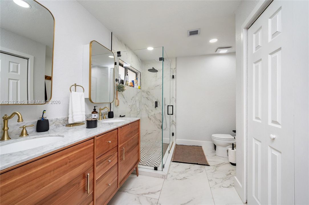Wood-finish double vanity with stone countertop, brass-tone fixtures, wall-mounted medicine cabinets, and glass-enclosed walk-in shower with stone-style wall tile and patterned floor tile