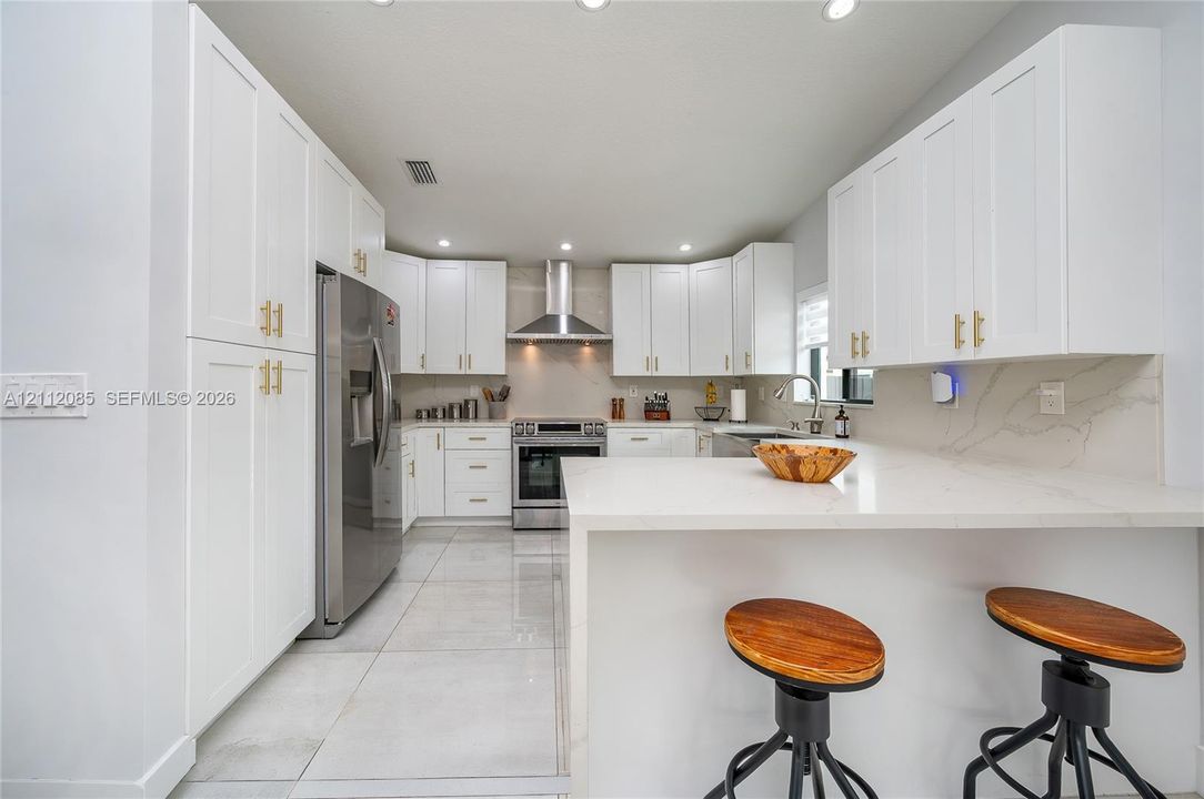 Kitchen featuring white shaker-style cabinetry, quartz countertops, stainless steel appliances, a stainless steel range hood, and large-format floor tiling