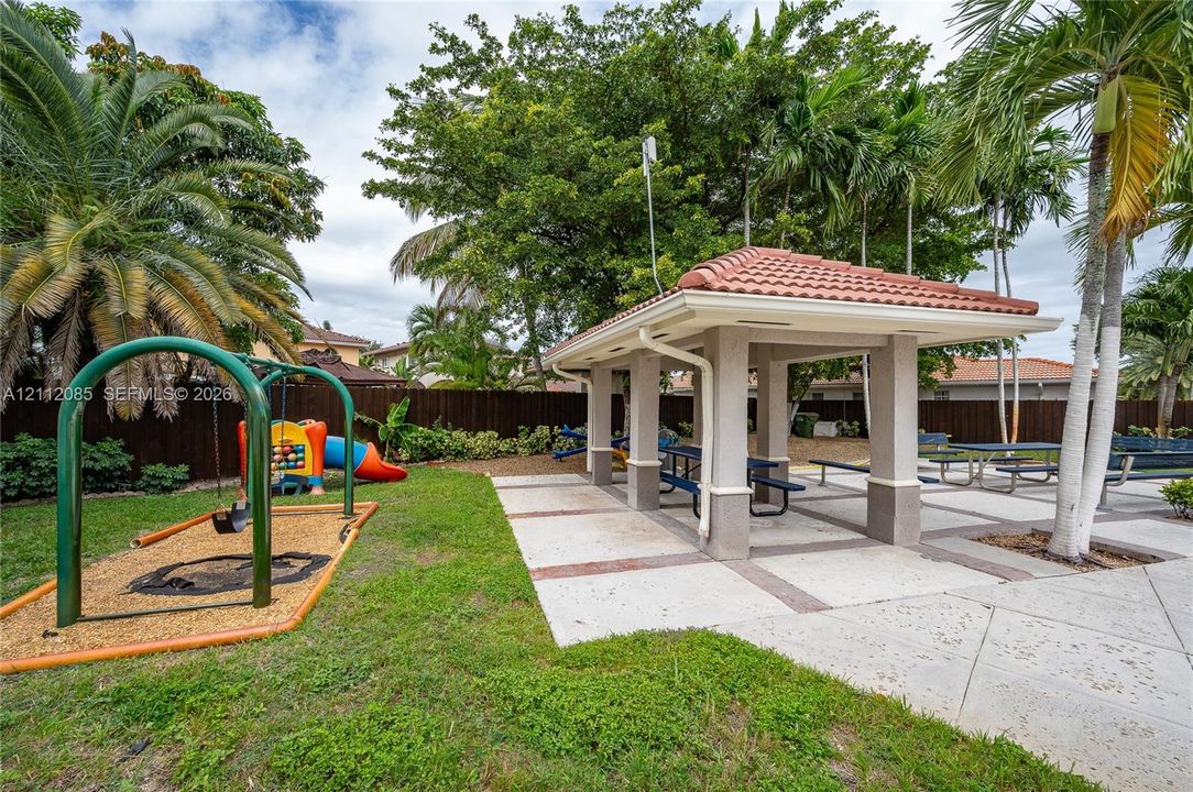 Outdoor recreation area featuring a covered pavilion with a tiled roof, concrete hardscaping, manicured lawn, and mature palm trees
