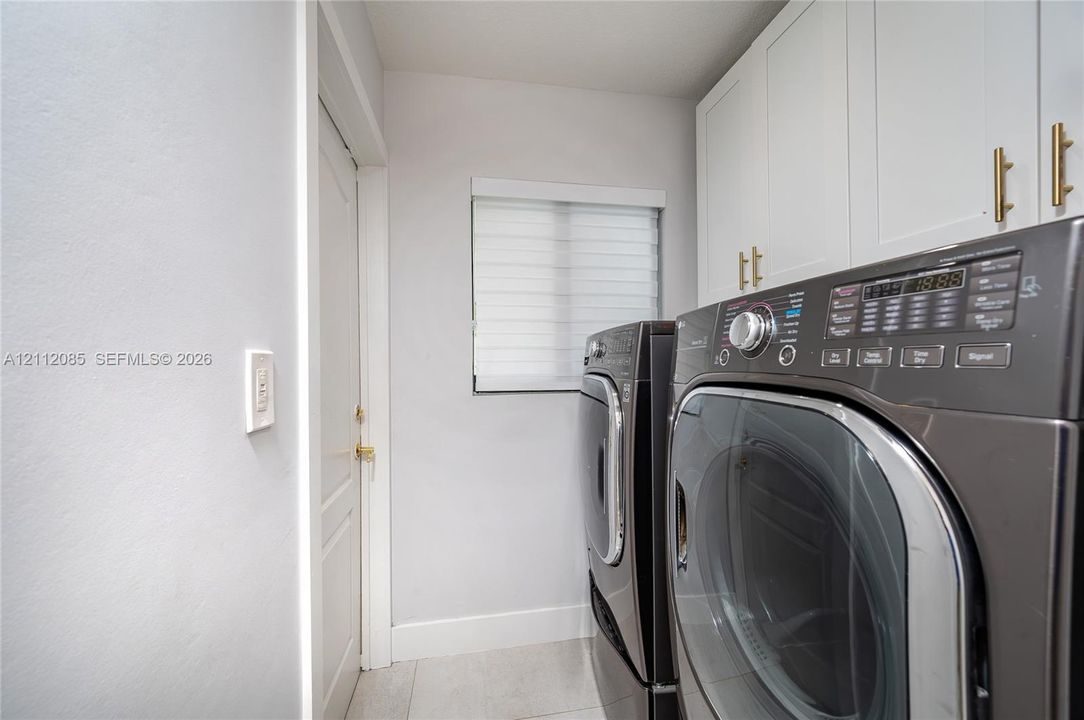 Utility space featuring white shaker-style wall cabinets with metallic hardware, tile flooring, and a window with horizontal blinds