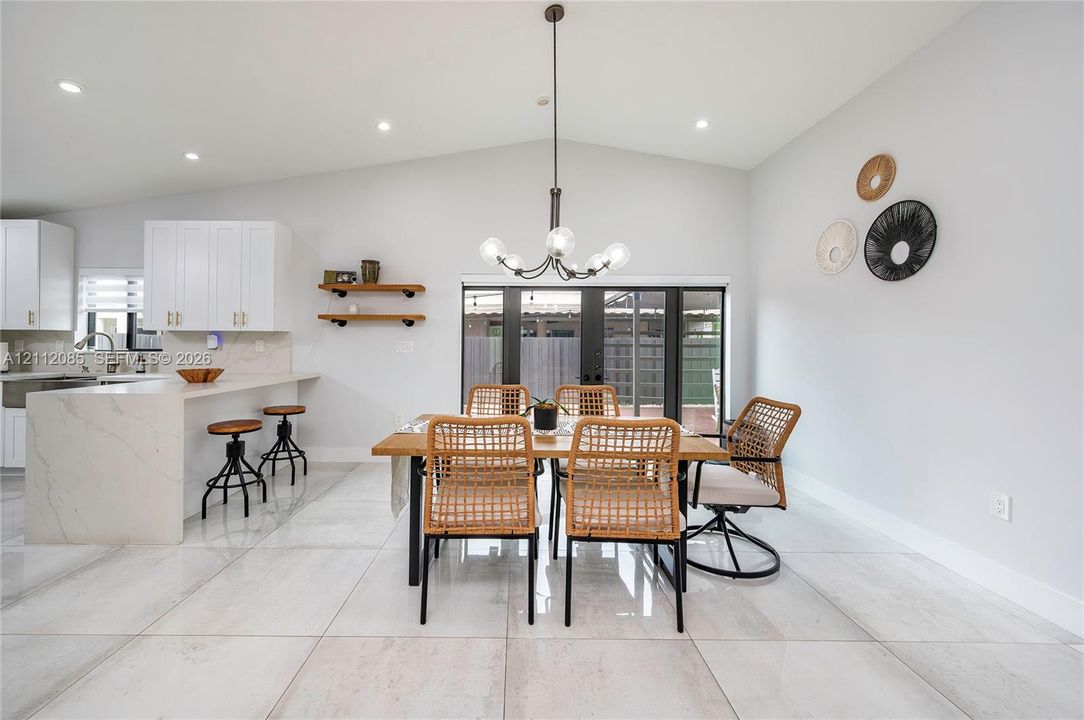 Open-concept interior featuring vaulted ceilings, oversized tile flooring, a kitchen peninsula with a stone-finish countertop, floating wood shelving, and a multi-light chandelier