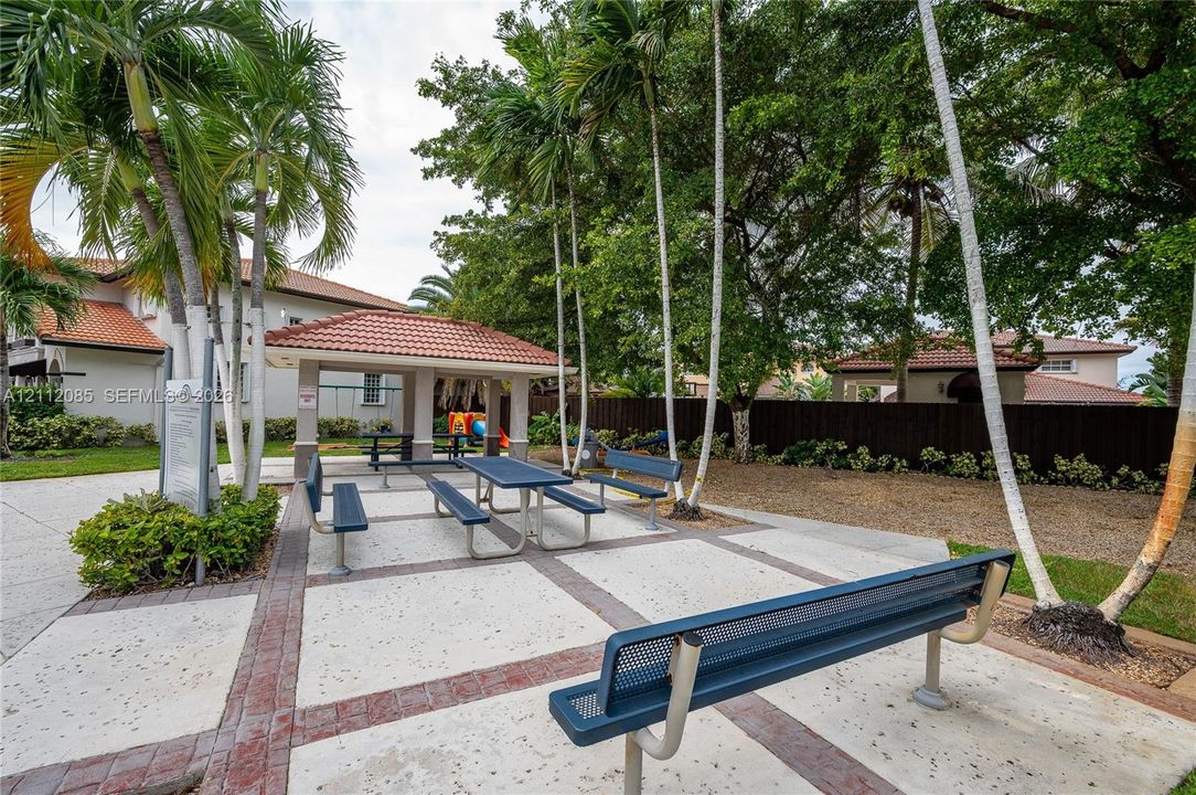 Covered pavilion with tile roof, concrete patio with masonry accents, metal park benches, and mature palm trees