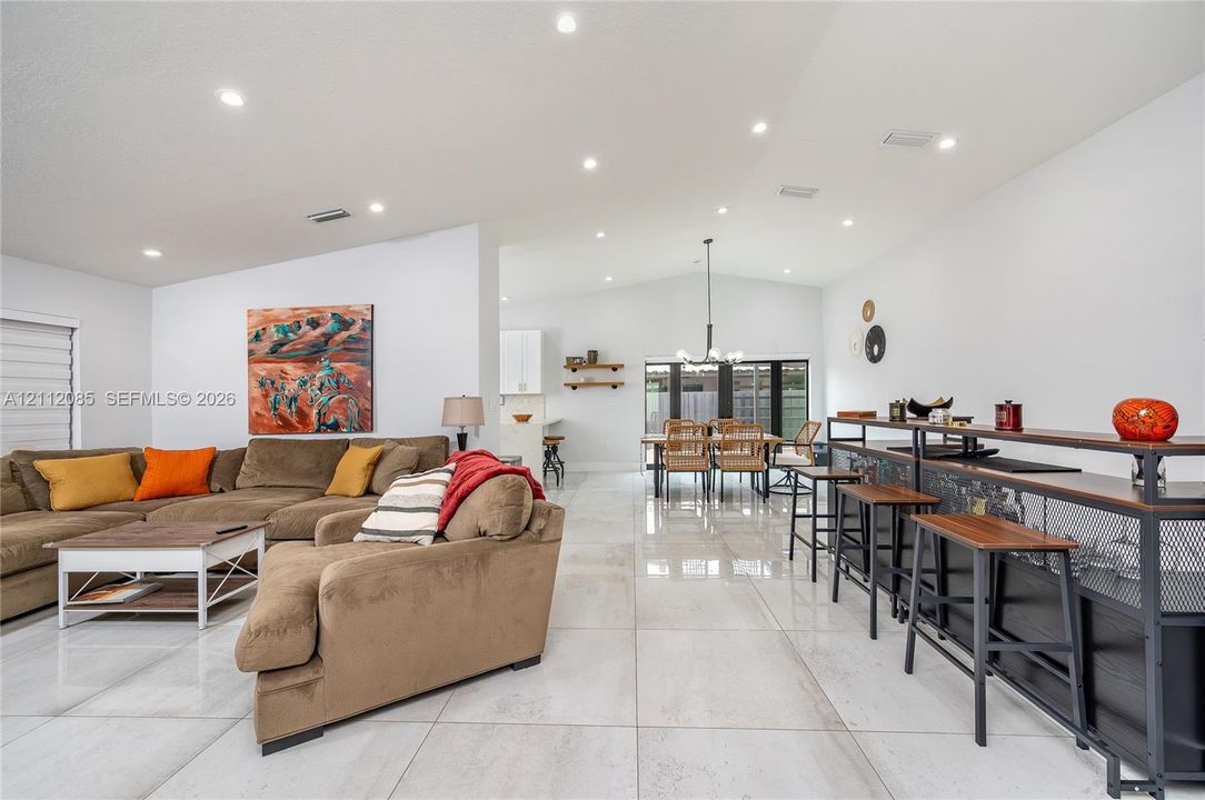 Open-concept area featuring large-format tile flooring, vaulted ceilings, recessed lighting, and a built-in wet bar with dark metal and stone-finish cabinetry