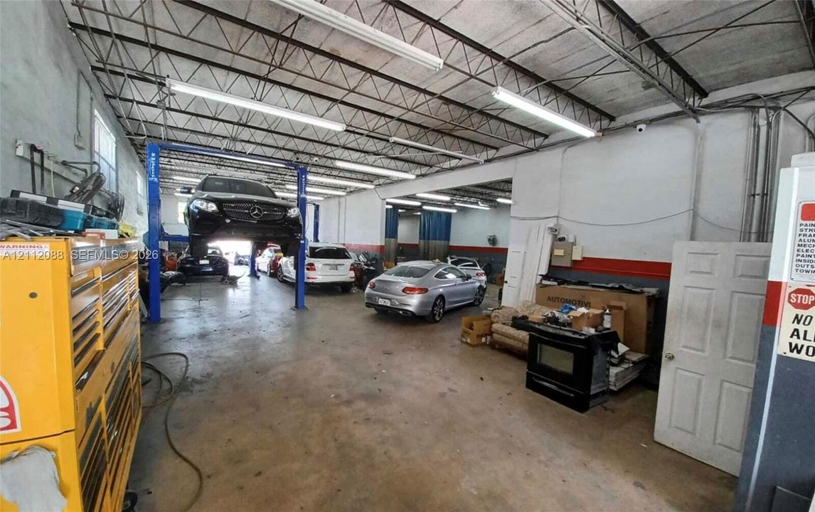 Spacious industrial workshop featuring high-clearance exposed steel truss ceilings, concrete flooring, integrated automotive lift system, and overhead fluorescent lighting