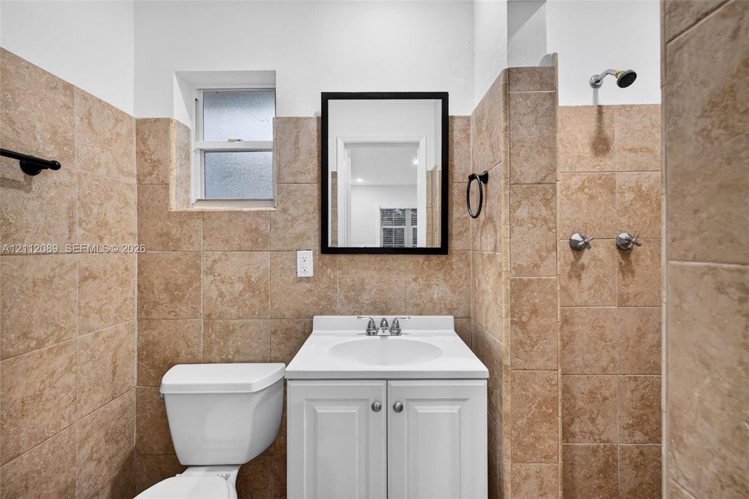 Bathroom featuring tan stone-tile wall cladding, a white pedestal vanity with an integrated sink, a framed wall mirror, and a wall-mounted shower head