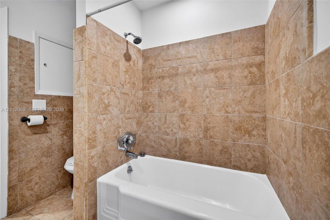 Full-height tan stone-finish tile surround, white acrylic soaking tub, wall-mounted showerhead, and integrated chrome faucet hardware
