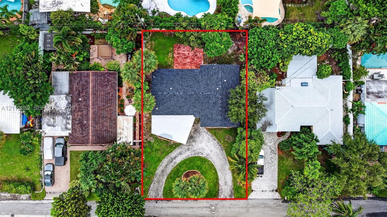 Aerial perspective of property featuring a dark-toned shingle roof, a circular driveway, mature perimeter landscaping, and a rear stone-paved patio