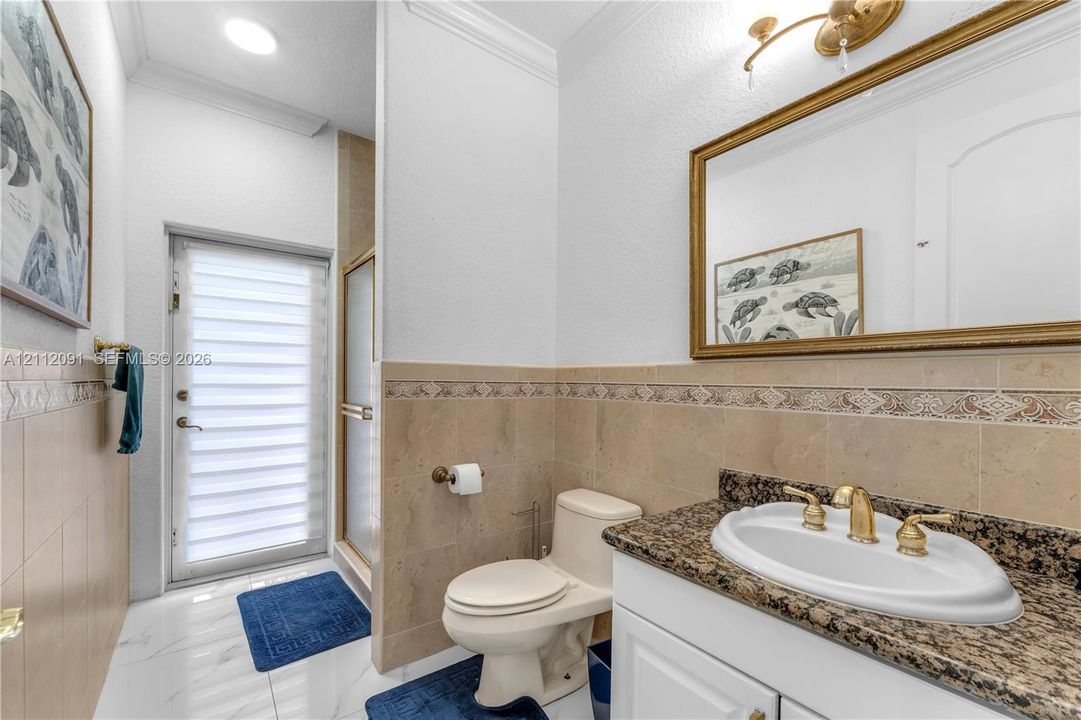 Bathroom featuring stone-finish countertop, undermount sink with gold-tone hardware, decorative tiled wall accent, glass-enclosed shower, and opaque-paned exterior door