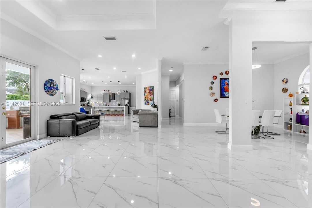 Open-concept main living area featuring large-format polished tile flooring, recessed lighting, tray ceilings, and a glass-paneled door leading to an outdoor space