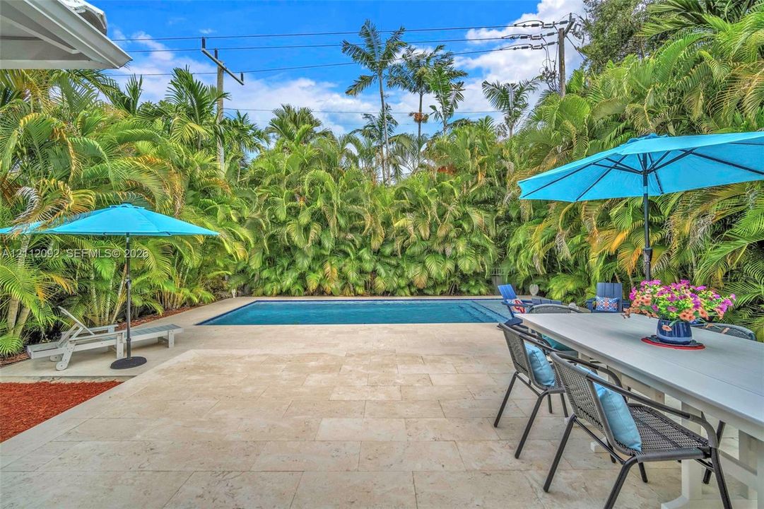 Rectangular swimming pool with stone-finish decking, surrounded by dense perimeter landscaping and mature palm trees