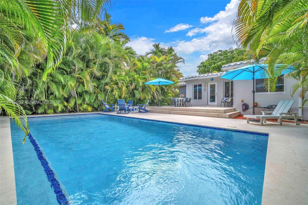 In-ground swimming pool with tile surround, concrete patio, and dense perimeter landscaping
