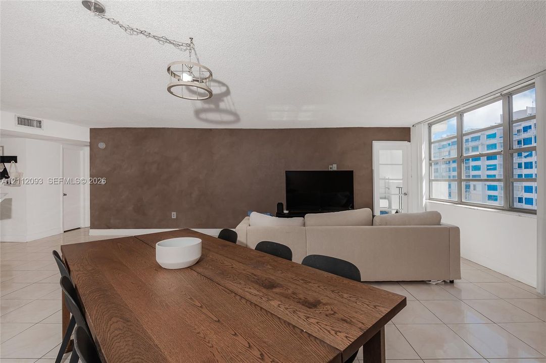 Open-concept living space featuring large-format ceramic tile flooring, a prominent accent wall with textured finish, expansive multi-pane windows, and a suspended industrial-style drum pendant light fixture