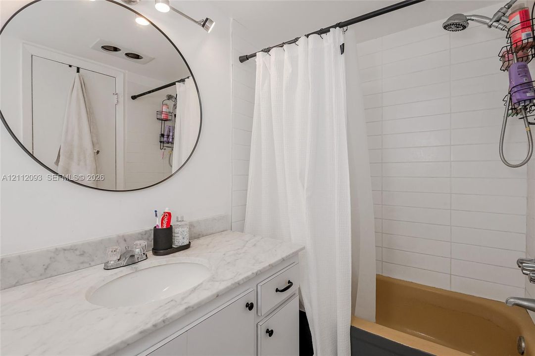Stone-topped vanity with an integrated oval sink, circular wall-mounted mirror, and white horizontal tile wall surround in the shower area