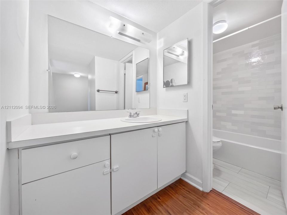 Bathroom featuring a vanity with a white countertop and integrated sink, large wall-mounted mirror, wood-finish flooring, and a bathtub with tile surround