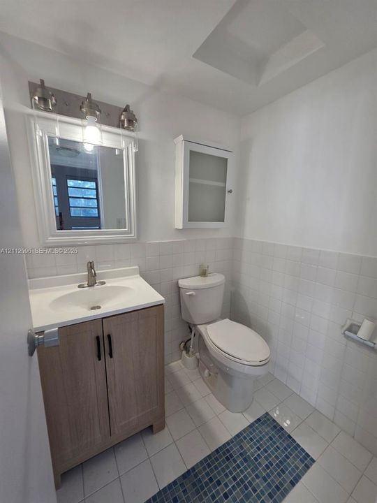 Bathroom featuring a wood-finish vanity with an integrated basin, a white-framed mirror with three-bulb vanity lighting, a wall-mounted glass-paneled medicine cabinet, a dual-flush toilet, and white tile flooring with a central blue mosaic inlay