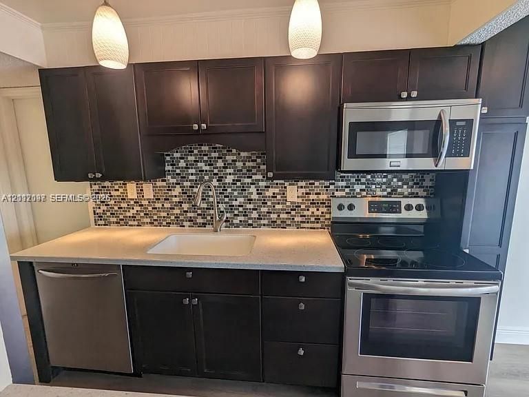 Dark-toned cabinetry paired with light solid-surface countertops, multi-toned mosaic tile backsplash, stainless steel appliances, and pendant lighting