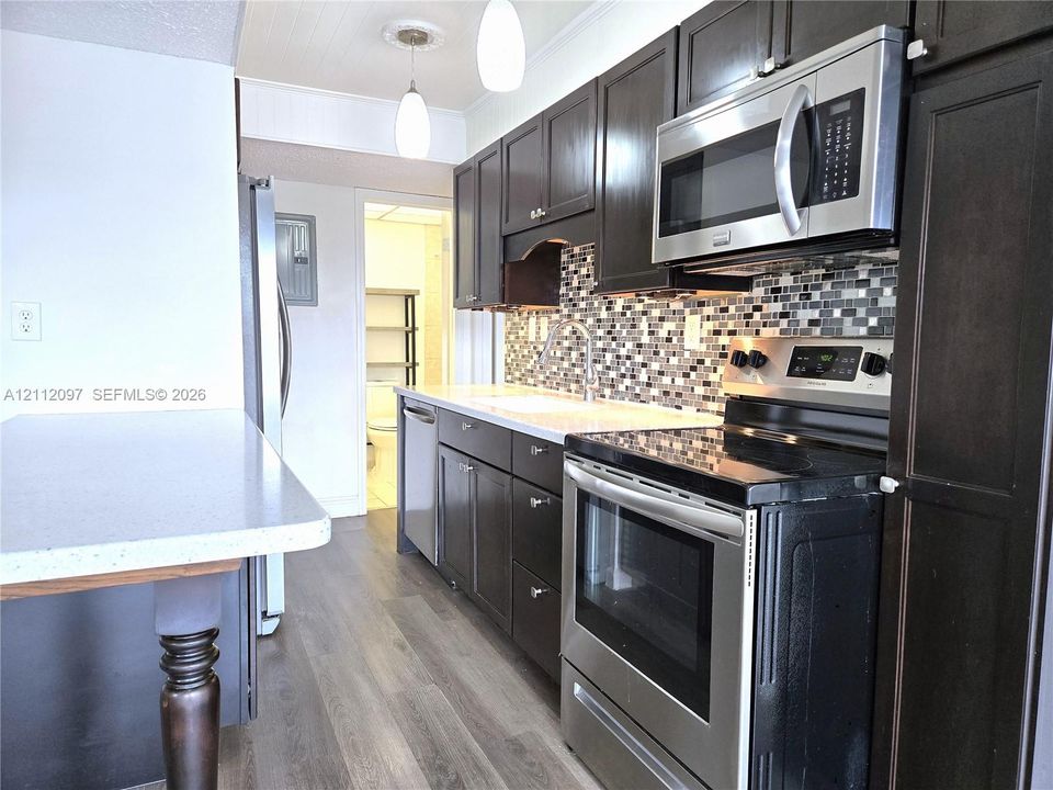 Kitchen featuring dark-finish cabinetry, stainless steel appliances, multi-toned mosaic tile backsplash, quartz countertops, and wood-finish flooring