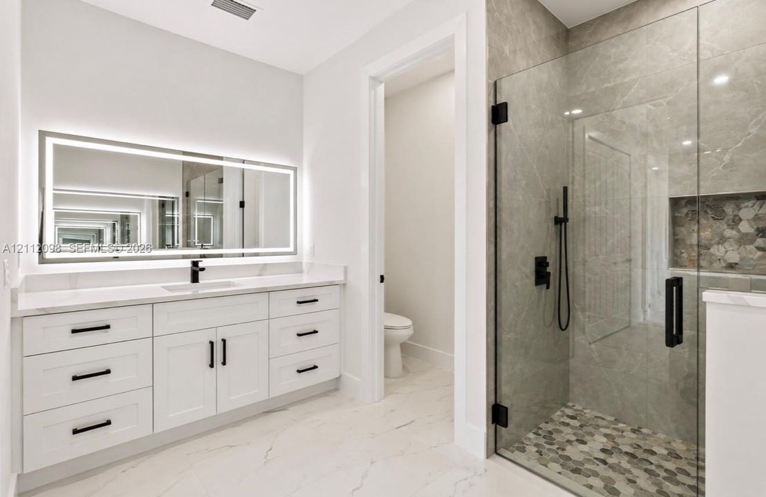 Bathroom featuring a white shaker-style vanity with solid-surface countertop, an integrated LED-backlit mirror, a walk-in shower with large-format tile surround, matte black hardware, and hexagonal tile flooring