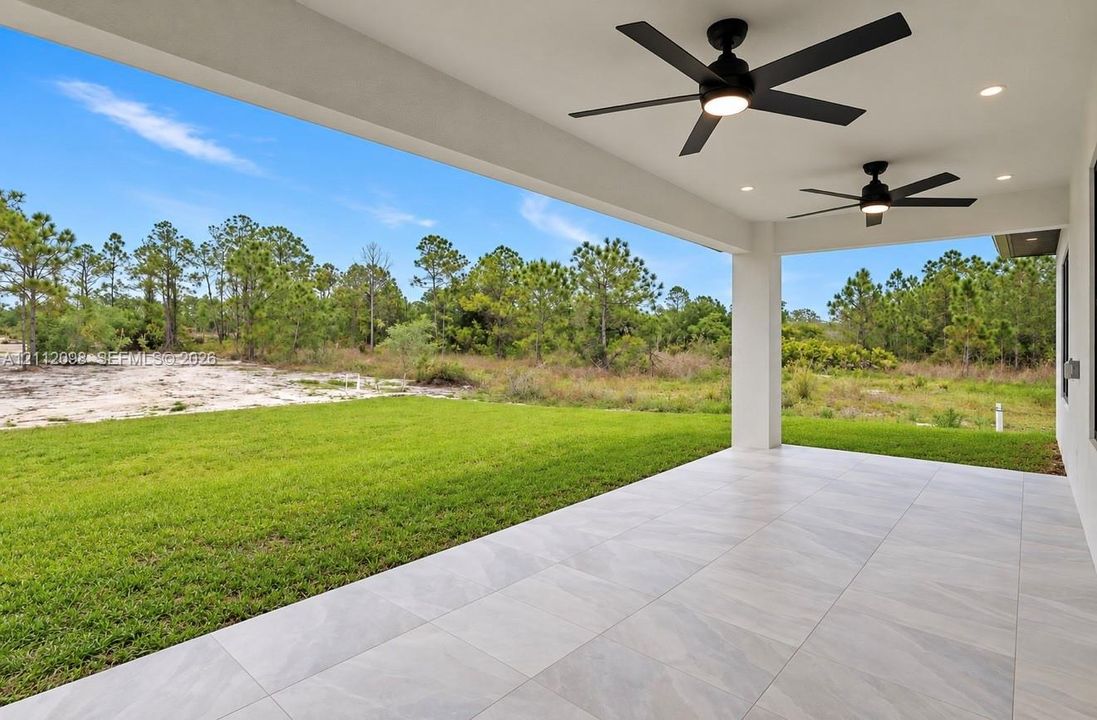 Covered patio featuring large-format porcelain tile flooring, dual ceiling fans with integrated lighting, recessed lighting, and a rear yard bordering natural vegetation