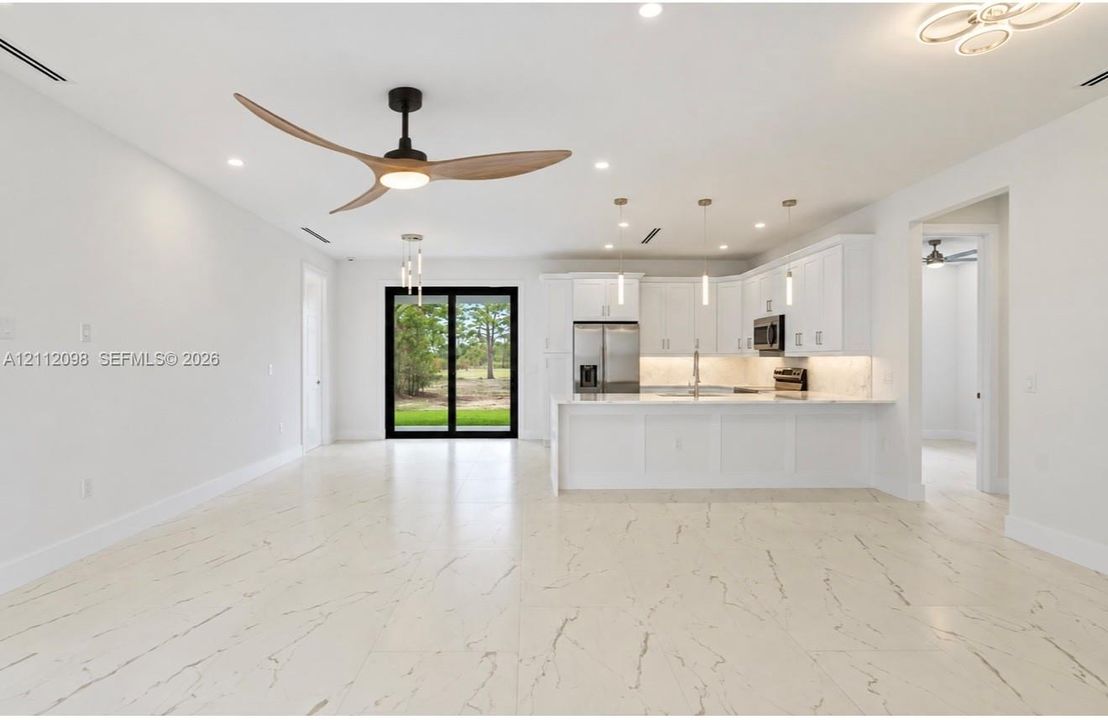 Open-concept area featuring white cabinetry with integrated stainless steel appliances, large kitchen island with light-toned stone-finish countertop, polished porcelain tile flooring, modern three-blade ceiling fan, and sliding glass doors
