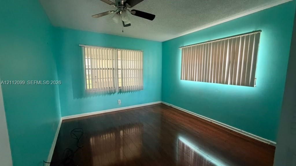 Room featuring wood-finish flooring, two sets of windows with vertical blinds, and a multi-blade ceiling fan with light fixture