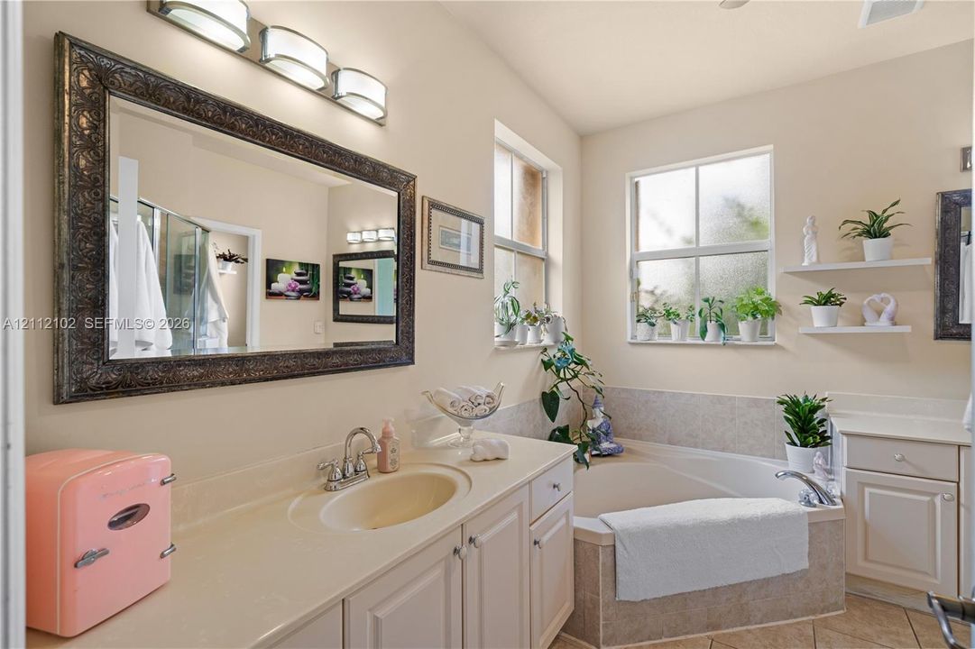 Bathroom featuring a vanity with integrated sink, large framed wall mirror, three-light fixture, corner soaking tub with tiled surround, and wall-mounted shelving