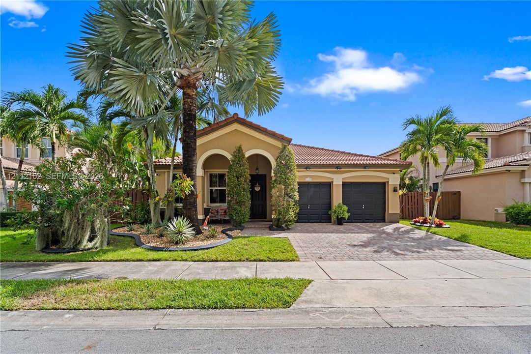 Single-story residence featuring a tile roof, dual-bay garage, arched entryway, and paver driveway surrounded by tropical landscaping