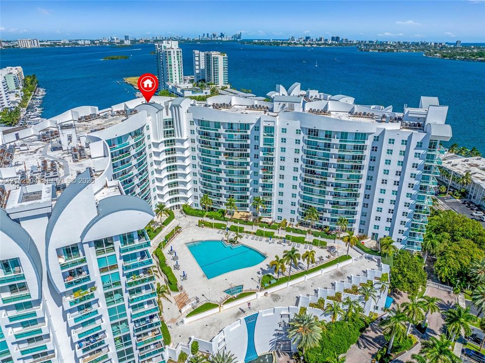 High-rise residential exterior featuring expansive waterfront positioning, glass-railed balconies, a central swimming pool, and tropical landscaping
