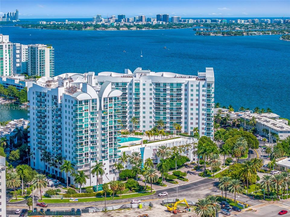 High-rise residential complex featuring a podium-level swimming pool, expansive glass-railed balconies, and extensive perimeter landscaping with mature palm trees