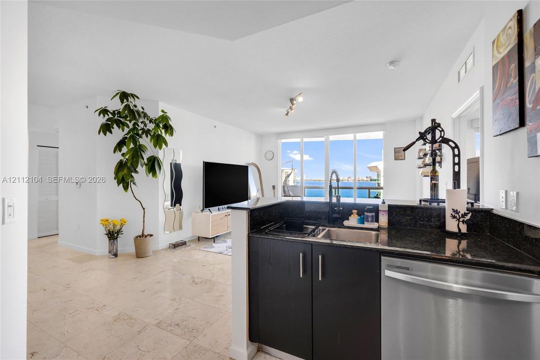 Kitchen featuring dark cabinetry, stone countertops, stainless steel dishwasher, and under-mount sink, complemented by large-format tile flooring and floor-to-ceiling windows