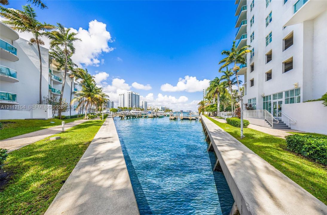 Waterfront canal bulkhead featuring concrete walkways, manicured landscaping, palm trees, and multi-story structures with glass-railing balconies