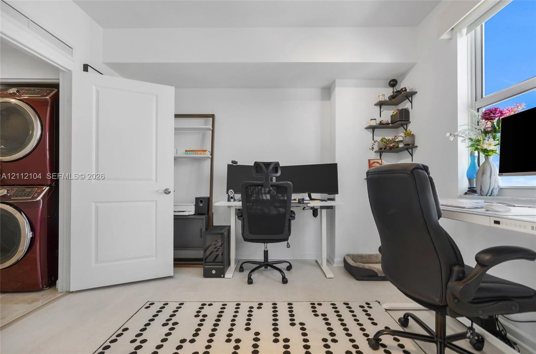 Room featuring neutral wall-to-wall carpeting, a large window, wall-mounted tiered shelving, and an enclosed laundry closet with stacked washer and dryer unit