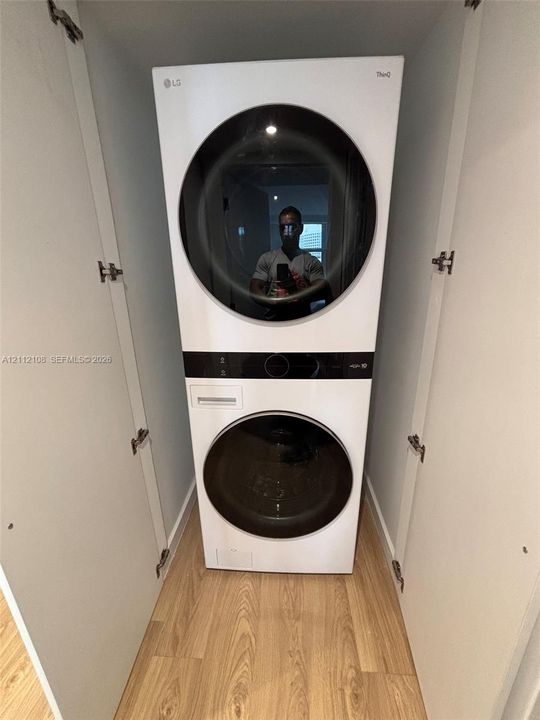 Stacked laundry unit featuring a white finish, circular glass-front doors, integrated digital control panels, and light-toned wood-finish flooring