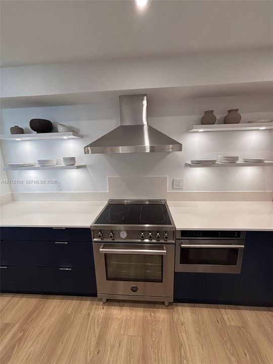 Kitchen featuring dark flat-panel cabinetry, quartz countertops, a stainless steel range with a matching chimney-style hood, and integrated under-shelf lighting
