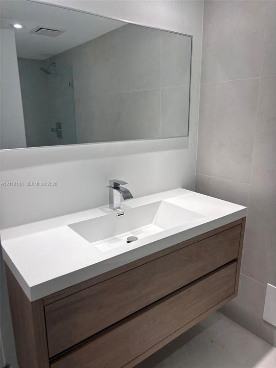Floating vanity featuring a wood-finish cabinet with dual drawers, integrated white sink, polished chrome faucet, and large wall-mounted mirror against a backdrop of neutral stone-finish wall tile