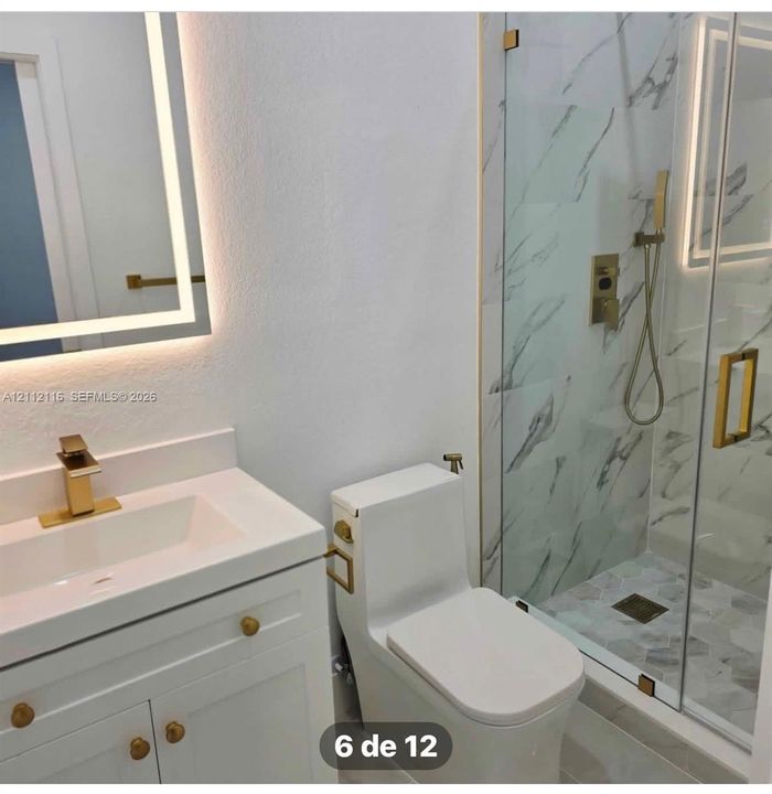 Bathroom featuring a white vanity cabinet with gold-tone hardware, a wall-mounted LED-backlit mirror, a seamless-glass shower enclosure with gold-tone fixtures, and patterned stone-finish shower wall tile